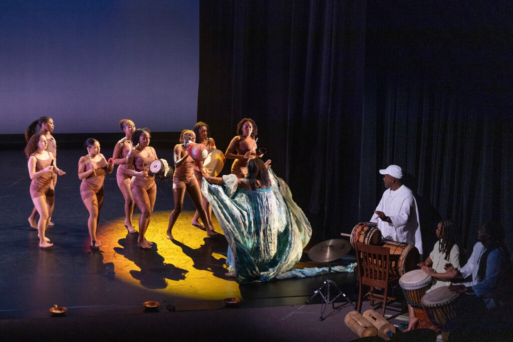 Dancers in spotlight, accompanying drummers at bottom right