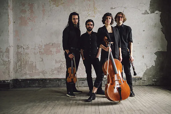 Musicians with instruments standing in a room with bare floors and cracked, peeling walls.