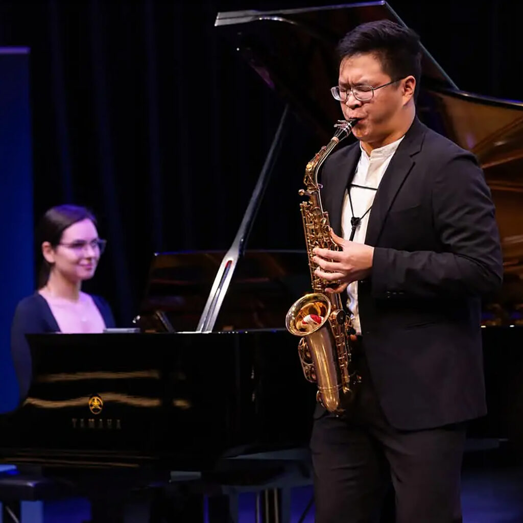 Performance photo of young man playing saxophone with accompanist in background