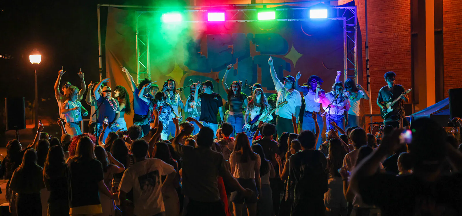 Performers and audience during nighttime concert, stage lit with colorful lights