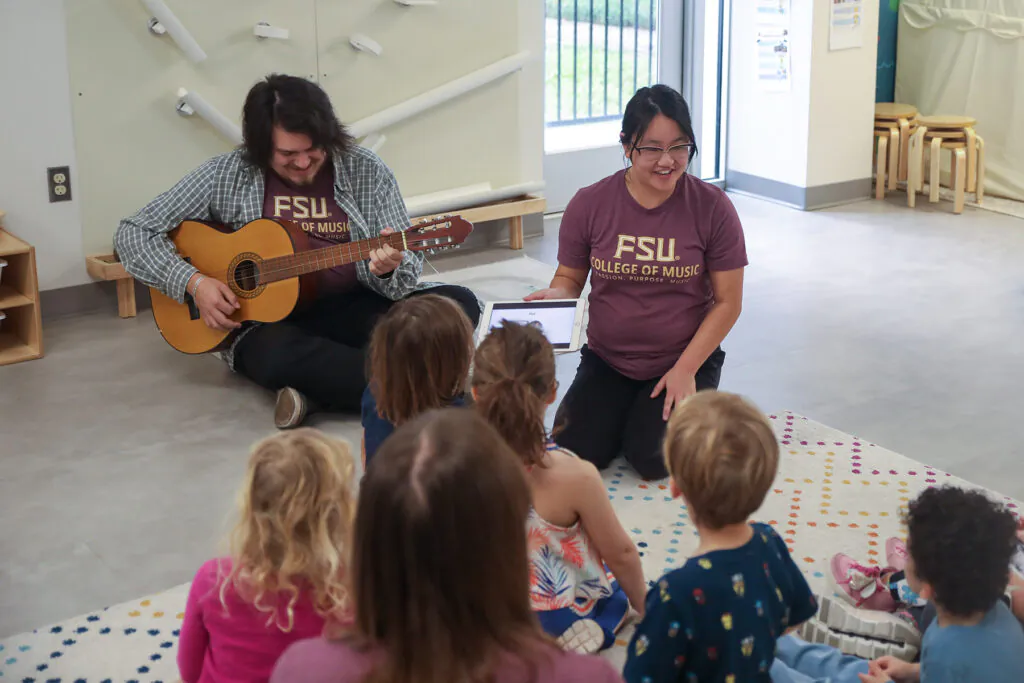 Young man with guitar and young woman with tablet interact with a group of children