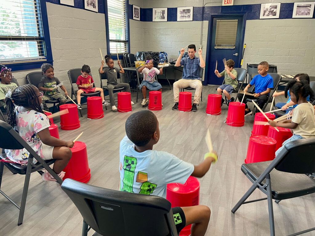 Young man leads a group of children as they hit plastic buckets with drum sticks
