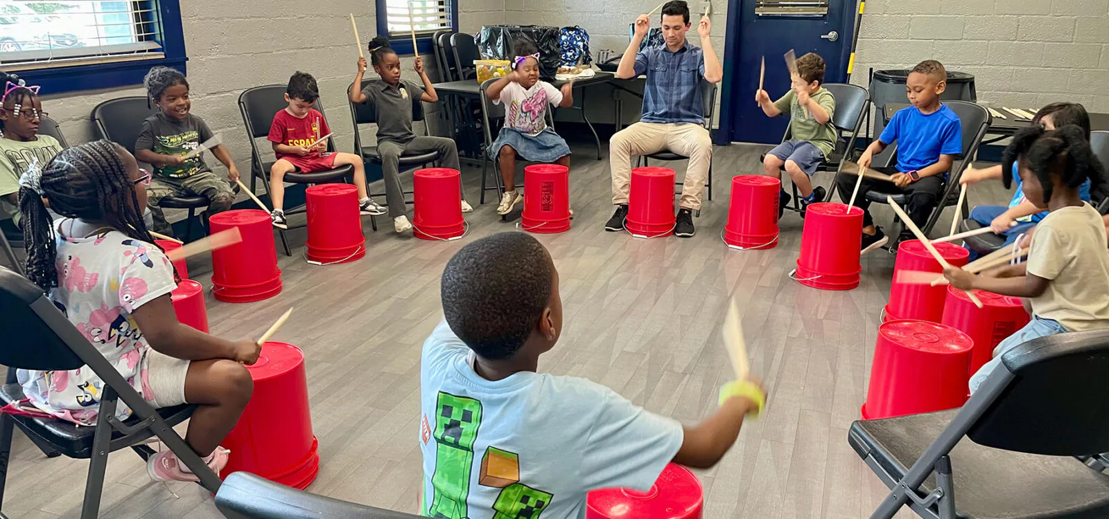 Young man leads a group of children as they hit plastic buckets with drum sticks