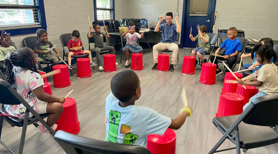 Young man leads a group of children as they hit plastic buckets with drum sticks