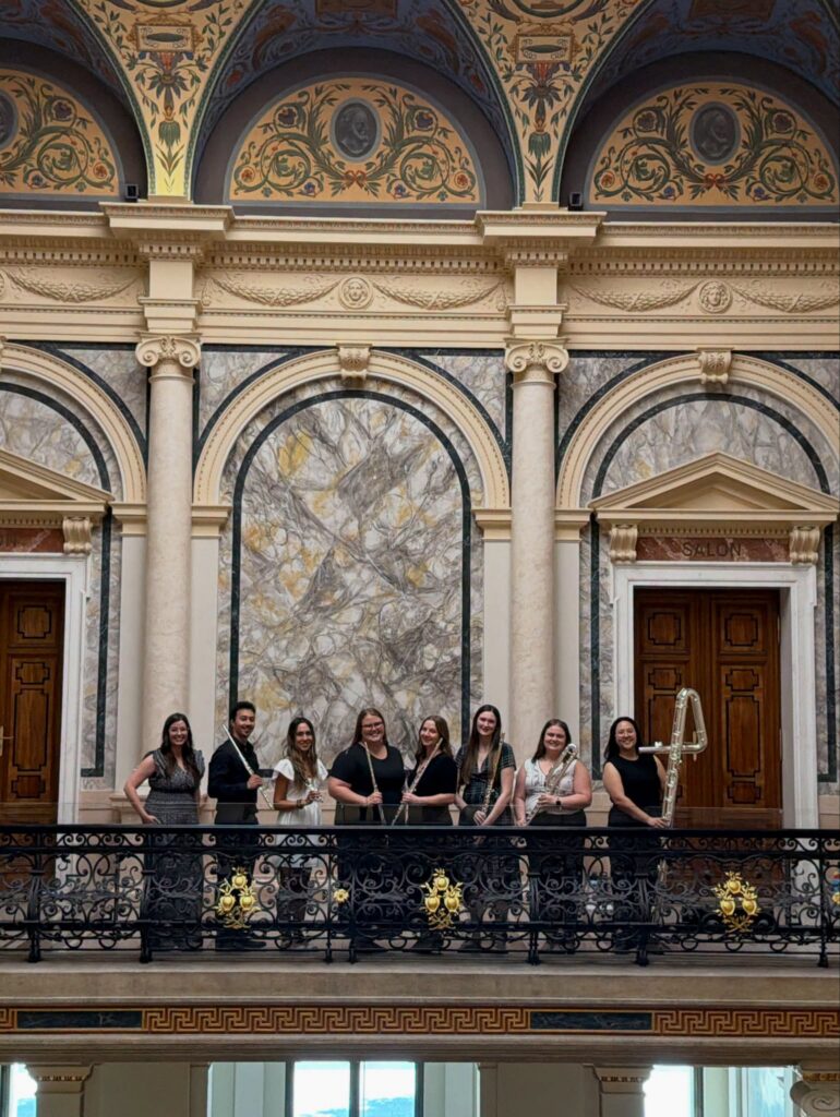 Group of students holding flutes pose with teacher on ornate balcony