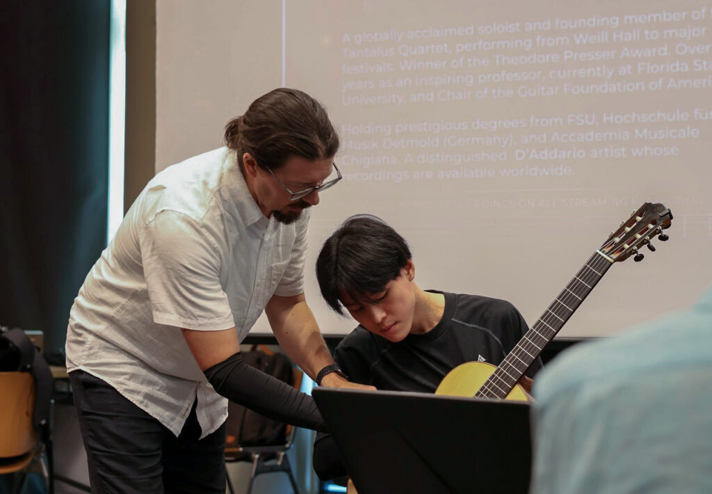 young man holding guitar while teacher points out feature of instrument