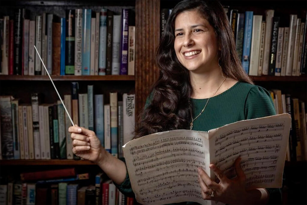 Smiling woman holding sheet music and baton posing in front of bookcase